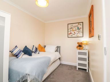A small bedroom with a single bed pillows and a blanket beside a white drawer with a teddy bear and lamp on top at Arnside House South Lakeland Leisure Village