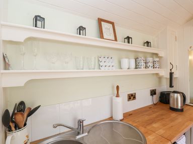 A kitchen countertop with a sink a paper towel holder a kettle and a toaster below shelves with glasses mugs and small lanterns at Woodlands Retreat Shepherd's Hut Brundish near Laxfield