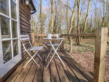 A wooden deck with two white metal chairs and a small round table with mugs and croissants in a wooded area at Woodlands Retreat Shepherd's Hut Brundish near Laxfield