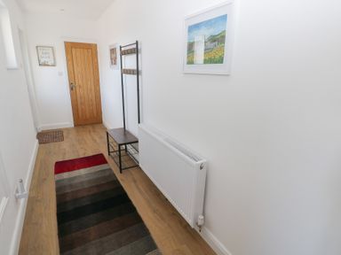 A narrow hallway with a wooden door a multi-colored rug a coat rack with a bench a radiator and framed pictures on white walls at Arfryn in Solva