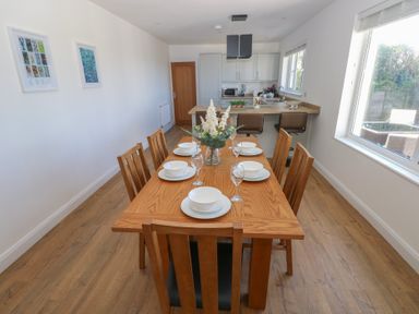 A dining room with a wooden table set for six with plates and glasses near a kitchen with bar stools at Arfryn in Solva