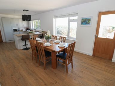 A dining area with wooden table set with plates and glasses adjacent to a kitchen with white cabinets and stools at Arfryn in Solva