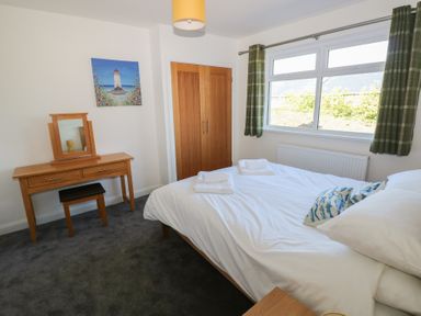 A bedroom with a bed, wooden vanity table with mirror and stool, a window with green curtains and a wooden closet at Arfryn in Solva