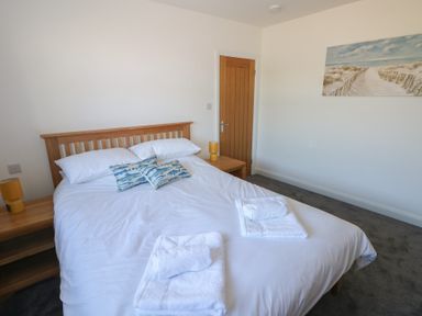 A bedroom with a double bed white linens towels wooden headboard side tables yellow lamps and a beach painting on the wall at Arfryn in Solva