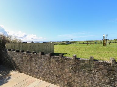 An outdoor area with a stone wall a wooden fence and a playground on grass at Arfryn in Solva