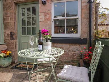 A small outdoor seating area with a metal table and chairs with cushions flowers on the table and against the house at Dromore Cottage in Norham