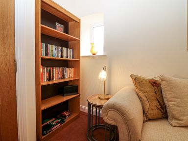A bookshelf with books and board games next to a window with a vase and a lamp on a side table by a sofa with cushions at Dromore Cottage in Norham