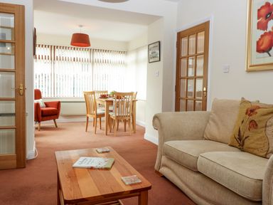 A living room with a beige sofa and wooden coffee table leading to a dining area with wooden table and chairs at Dromore Cottage in Norham
