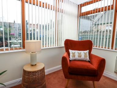 A corner with a red armchair with a cushion and a wicker side table with a lamp next to windows with vertical blinds at Dromore Cottage in Norham