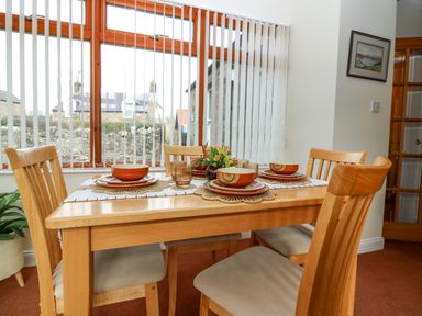 A dining area with a wooden table set for four with orange bowls and plates near a window with vertical blinds at Dromore Cottage in Norham