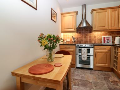 A kitchen with wooden cabinets and tiled backsplash a stainless steel oven a wooden table with a vase of flowers and circular wooden placemats at Dromore Cottage in Norham