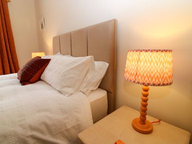 A bedroom with a beige upholstered headboard white bedding an orange pillow an orange lamp on a wooden bedside table and orange curtains at Dromore Cottage in Norham