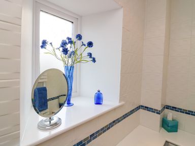 A bathroom window shelf with a small round mirror a blue vase with flowers a blue bottle and a blue tissue box at Dromore Cottage in Norham
