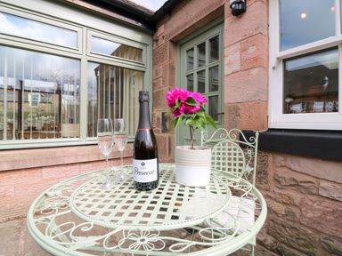 A metal patio table with a bottle of prosecco two glasses and a potted flower outside a stone building at Dromore Cottage in Norham