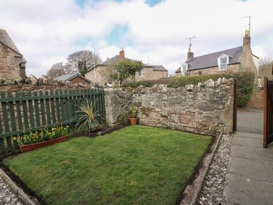 A small enclosed garden with grass a stone wall a green wooden fence and flowers at Dromore Cottage in Norham