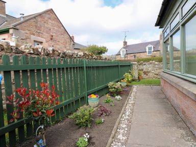 A garden area with a green wooden fence planted flowers and stone paved path at Dromore Cottage in Norham