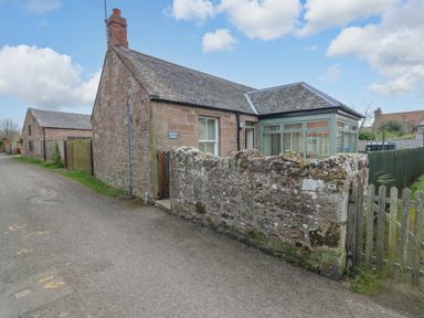 An exterior view of a stone cottage with a small stone wall and wooden gate along a road at Dromore Cottage in Norham