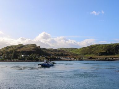 A boat on water with hills and houses in the background at WildLuing Cullipool Isle Of Luing