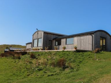 A wooden cabin with large windows and flower pots on a deck in a grassy area at WildLuing in Cullipool Isle Of Luing