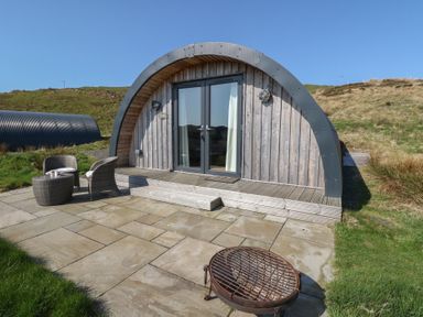 An arched wooden cabin with glass doors a small patio with two chairs a table and a fire pit at WildLuing Cullipool Isle Of Luing