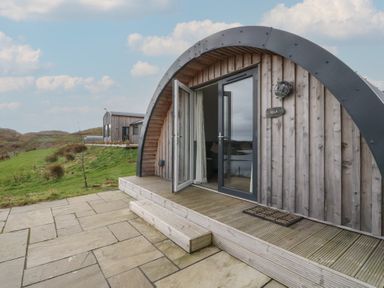 An arched wooden cabin with a glass door open onto a stone patio and grassy landscape at WildLuing Cullipool Isle Of Luing