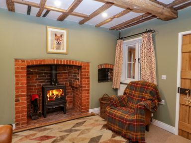 A room with a brick fireplace with a wood stove fire armchair with a plaid blanket window with curtains and wooden ceiling beams at 2 Loxter Villas in Wellington Heath near Ledbury