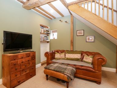 A living room with a brown leather sofa with cushions a wooden chest of drawers with a TV and bookshelves under exposed wooden beams at 2 Loxter Villas Wellington Heath near Ledbury