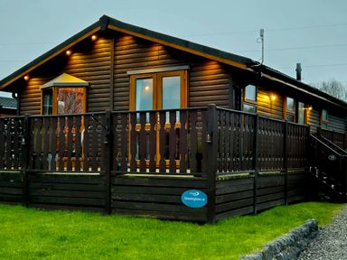 A wooden cabin with balcony railing and exterior lights on a grass lawn at Sunset Retreat in South Lakeland Leisure Village near Carnforth