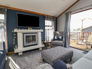 A living room with a fireplace a mounted TV a patterned armchair and ottoman and patio doors leading outside at Sunset Retreat in South Lakeland Leisure Village near Carnforth