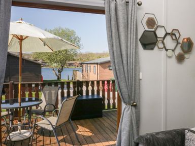A balcony with a table umbrella and chairs overlooking a lake with wooden cabins at Sunset Retreat in South Lakeland Leisure Village near Carnforth