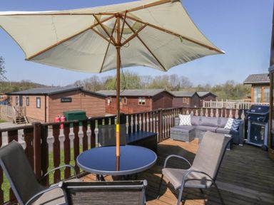 A wooden deck with outdoor table and chairs under an umbrella and a barbecue grill at Sunset Retreat South Lakeland Leisure Village near Carnforth