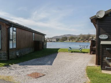 A gravel driveway between wooden buildings with a picnic table and chairs near a lake at Sunset Retreat in South Lakeland Leisure Village near Carnforth