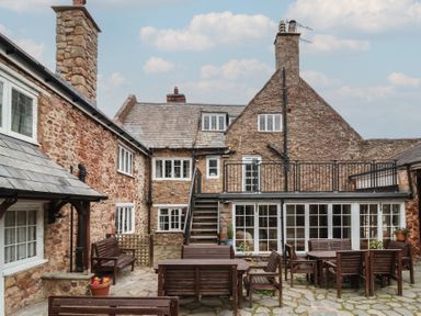 An outdoor stone courtyard with wooden tables and benches in front of a stone building at Dragon House in Bilbrook near Minehead