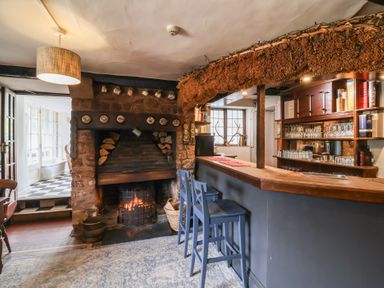A small bar area with wooden stools and a fireplace with stacked firewood at Dragon House in Bilbrook near Minehead