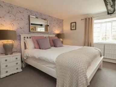 A bedroom with a white bed frame, pink and purple pillows, two lamps on nightstands, a window with beige curtains and floral wallpaper behind the bed at Dragon House in Bilbrook near Minehead