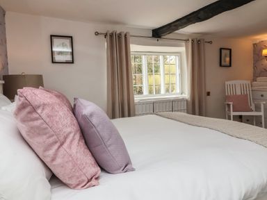 A bedroom with a bed featuring pink and purple pillows a window with curtains and a white chair with a pink cushion at Dragon House in Bilbrook near Minehead