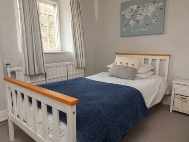 A bedroom with a white bed with blue blanket and pillows next to a window with striped curtains at Dragon House in Bilbrook near Minehead