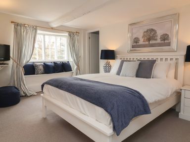 A bedroom with a white bed blue blanket and pillows window with curtains and cushions at Dragon House in Bilbrook near Minehead