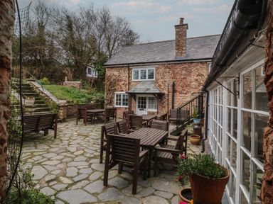 An outdoor patio with wooden tables and chairs stone paving and a stone house with a chimney at Dragon House in Bilbrook near Minehead