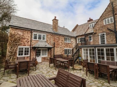 An outdoor patio area with wooden tables and benches in front of stone buildings with slate roofs at Dragon House in Bilbrook near Minehead