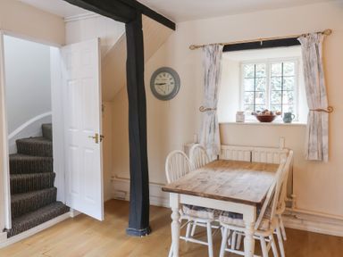 A dining area with a wooden table and four white chairs near a window with curtains and a clock on the wall at Dragon House in Bilbrook near Minehead