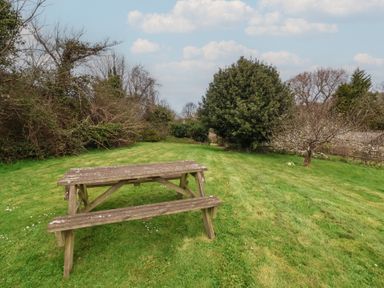 A wooden picnic table on a grassy lawn with trees and bushes in the background at Dragon House Bilbrook near Minehead