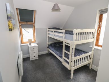 A bedroom with a white bunk bed blue bedding a small white bedside table with a lamp two windows and a wall-mounted heater at C65 Cahermore Holiday Village in Enniscrone County Sligo