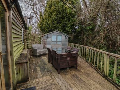 An outdoor wooden deck with a glass table and wicker chairs next to a wooden shed surrounded by trees at Gatekeeper's Cottage in Bilbrook near Minehead
