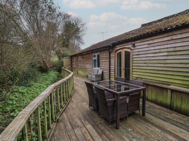 A wooden deck with a glass table and six chairs next to a wooden house with trees in the background at Gatekeeper's Cottage in Bilbrook near Minehead