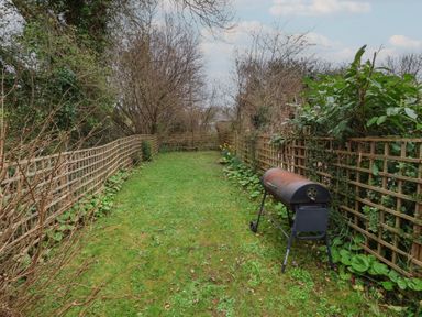 A narrow garden with grass lawn fenced on both sides and a barbecue grill at Gatekeeper's Cottage in Bilbrook near Minehead