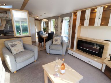 A living room with armchairs a coffee table with a bottle and glasses a wall unit and dining table at Lodge 2 in Silverdale