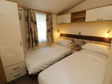 A bedroom with two single beds a wardrobe a window with curtains and overhead cabinets at Lodge 2 in Silverdale
