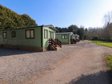 A row of green static caravans with steps on a gravel area beside a paved road with trees in the background at Lodge 2 in Silverdale