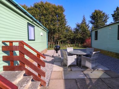 An outdoor patio area with a round table and four chairs between two light green lodges surrounded by trees at Lodge 3 in Silverdale